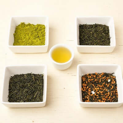 four white dishes full of green teas sit on a wooden surface with a single white cup of brewed green tea in center