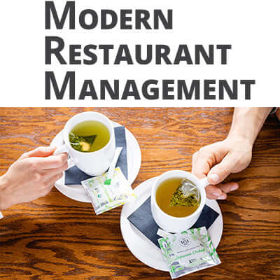 two hands grip white mugs with sachets brewing in them next to packaing that reads the tea spot on a wooden surface