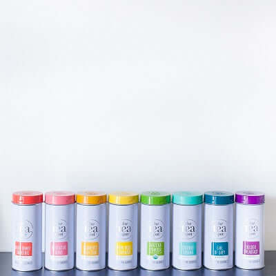 a line of tea tins with colorful labels sits on a countertop on a white background