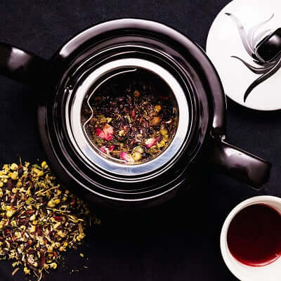 a teapot sits on a black surface next to its lid, a white cup of brewed tea and loose leaf tea blend