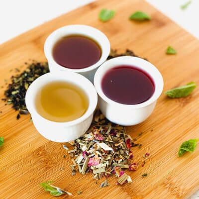 three cups of brewed tea sit on a bamboo surface next to mint leaves and loose tea