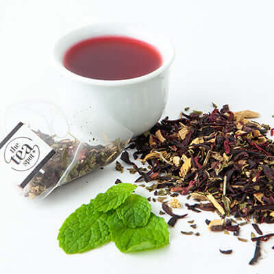 a cup of brewed tea sits next to a tea sachet, mint leaves and a pile of loose hibiscus tea
