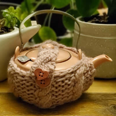 a teapot in a knitted cozy sits on a wooden surface next to potted plants