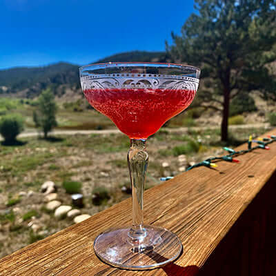a hibiscus tea cocktail sits on a wooden patio railling with a mountan view in the background