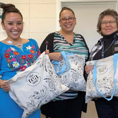 three women smile and hold tote bags with the tea spot logo on them