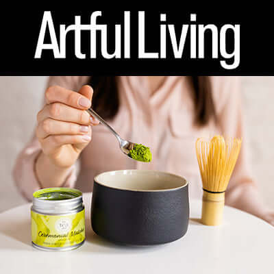 a person spoons matcha into a bowl on a table sitting next to a tin of ceremonial matcha and a matcha whisk