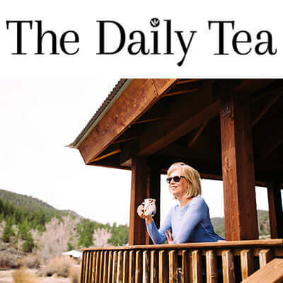 a woman stands on a cabin porch with roof smiling and holding a cup of tea in the mountains