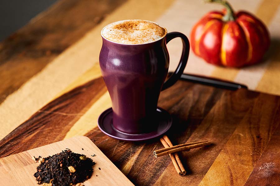 a wooden table with a tea latte in a mug sitting next to cinnamon sticks and a pile of tea with a pumpkin in the background