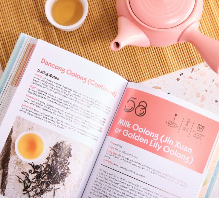 An open book 101 teas to steep before you die lies on a woven mat, with a pink teapot and two small cups of tea nearby. The pages show tea tasting notes and headings including 'Dancong Oolong (Continued)' and 'Milk Oolong (Jin Xuan or Golden Lily Oolong)'.