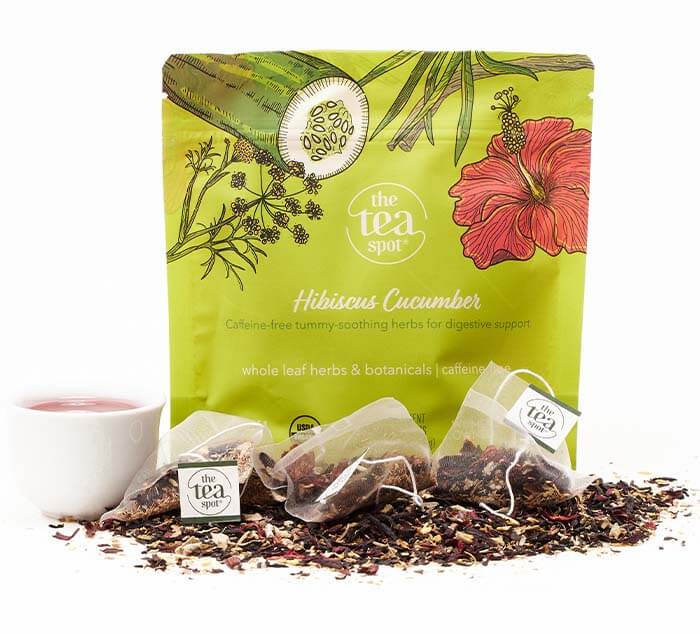 hibiscus cucumber herbal tes sachets sit on a pile of loose tea in front of a green floral pouch and cup of tea