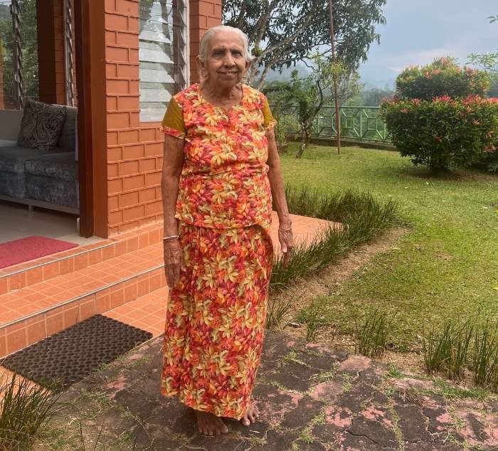 Mary Jayawardana in a floral dress standing on a stone path with a house and tea garden in the background