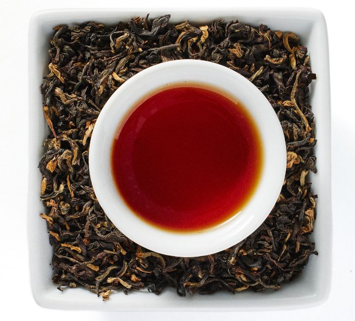 A top-down view of meleng assam loose leaf tea leaves arranged in a square white dish, with a small white bowl filled with brewed black assam tea in the center.