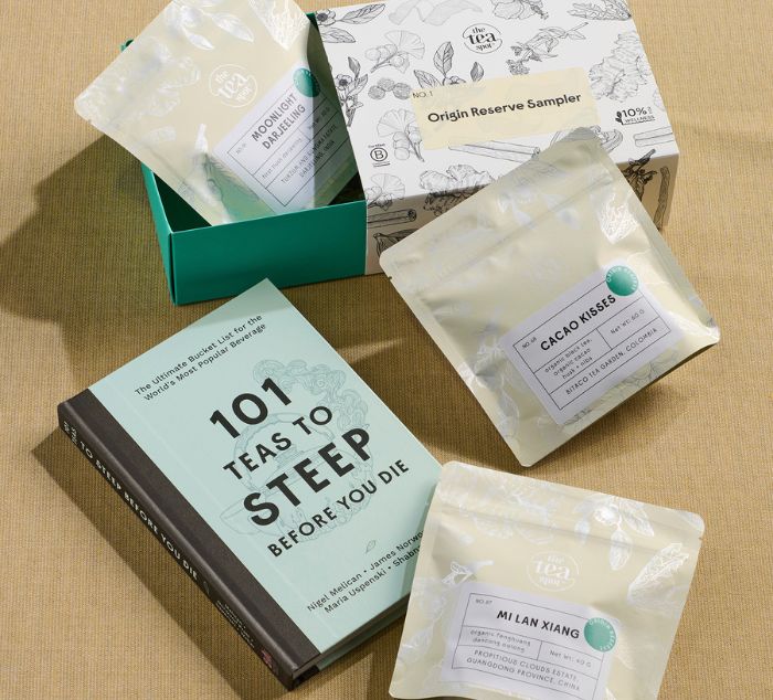 three shiny bags of tea sit with a gift box labeled 'Origin Reserve Sampler' and a book titled '101 Teas to STeep Before You Die' on a tan surface