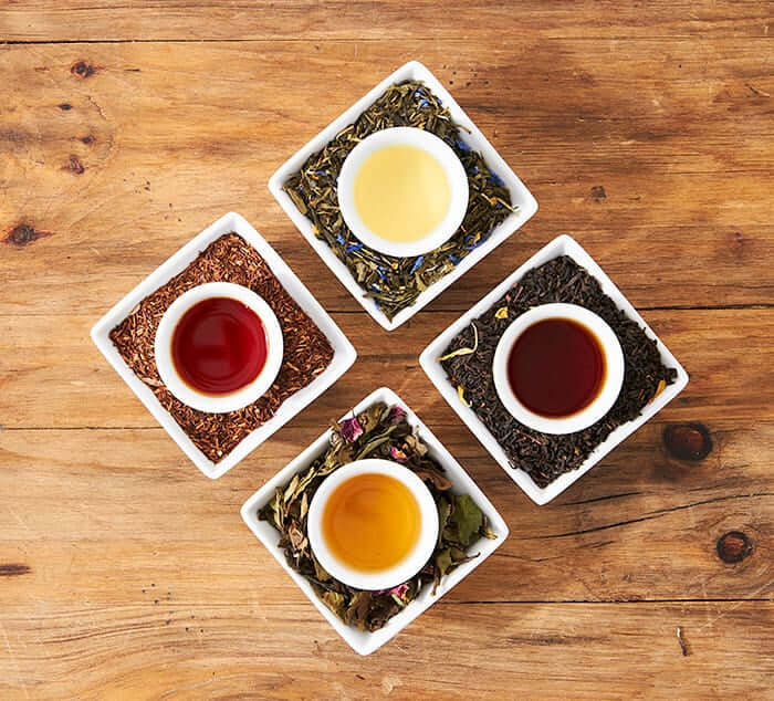 four cups of  brewed tea sit in dishes of loose tea blend on a wooden surface
